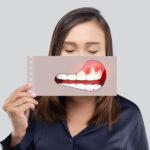 An Asian woman in a dark blue shirt holds a paper with an illustration of periodontal disease in front of her mouth, highlighting gum health issues.